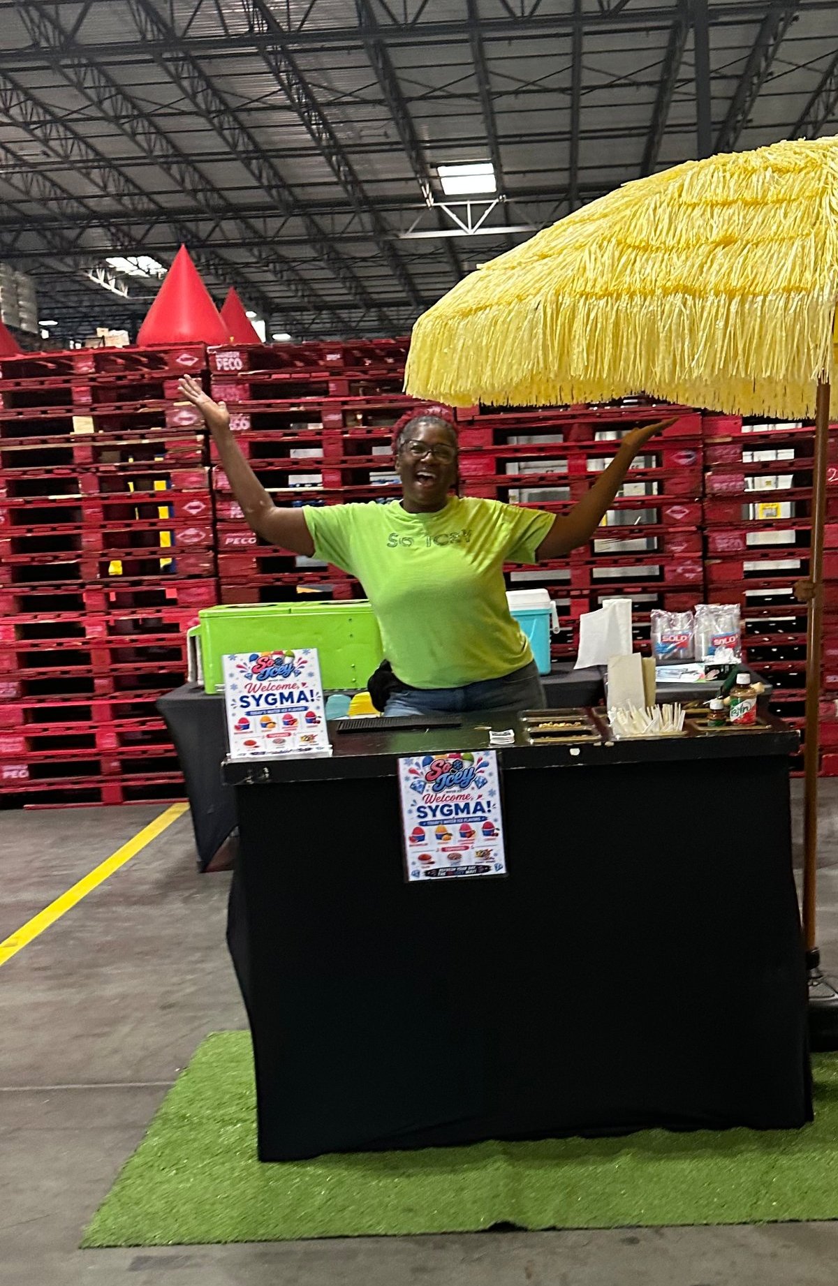 The So Icey founder at her setup under the yellow tiki umbrella, arms out mid-welcome at a warehouse event